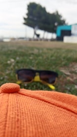 An orange hat is placed on grass with a pair of sunglasses in the background. The scene has trees and a building further behind, slightly out of focus.