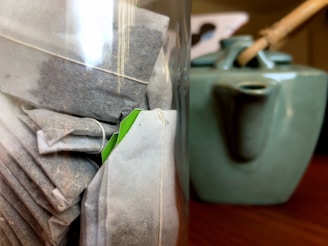 Several tea bags with tags are visible inside a glass jar, closely packed together. In the background, a ceramic teapot with a wooden handle is partially visible.