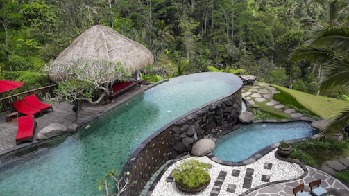 A luxurious outdoor infinity pool blends seamlessly into a dense tropical forest. The pool is flanked by a thatched-roof cabana with vibrant red lounge chairs on a wooden deck. Stone steps lead down to a smaller adjacent pool, and the area is adorned with lush greenery and palm trees.