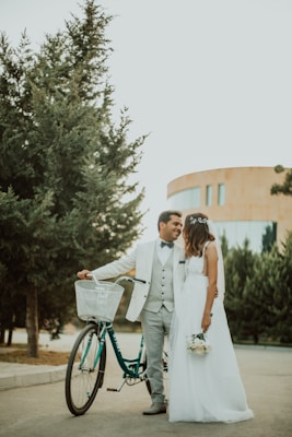 A couple stands in an outdoor setting, with the man in a white suit and the woman in a white wedding dress holding a small bouquet of flowers. They are close together, sharing a tender moment, and there is a vintage-style bicycle nearby. The background features lush green trees and a modern building, indicating a peaceful, possibly urban setting.