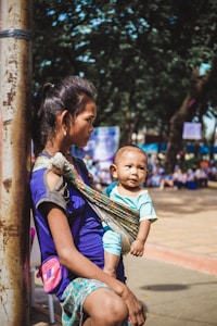 A woman is leaning against a pole while carrying a baby in a traditional sling. The baby is sitting comfortably in the sling, looking around with curiosity. The background shows a blurred scene of outdoors with people possibly sitting or gathering.