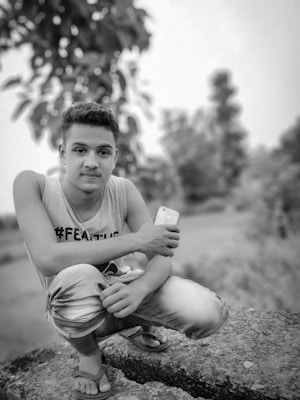 A young person is seen crouching outdoors on a rocky surface. They are wearing a sleeveless shirt with a hashtag printed on it and jeans. In one hand, they hold a smartphone. The photograph is in black and white, with blurred trees and foliage in the background, suggesting a natural environment.