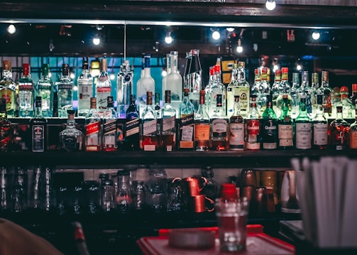 A well-stocked bar shelf filled with various bottles of liquor and spirits, including whiskey, vodka, gin, and rum. The bottles are lined up neatly on mirrored shelves, reflecting soft lighting from small bulbs above. Below the bottles, a row of upside-down glasses can be seen. There are colorful labels on the bottles, adding vibrancy to the scene.