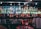 A well-stocked bar shelf filled with various bottles of liquor and spirits, including whiskey, vodka, gin, and rum. The bottles are lined up neatly on mirrored shelves, reflecting soft lighting from small bulbs above. Below the bottles, a row of upside-down glasses can be seen. There are colorful labels on the bottles, adding vibrancy to the scene.