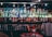 A well-stocked bar shelf filled with various bottles of liquor and spirits, including whiskey, vodka, gin, and rum. The bottles are lined up neatly on mirrored shelves, reflecting soft lighting from small bulbs above. Below the bottles, a row of upside-down glasses can be seen. There are colorful labels on the bottles, adding vibrancy to the scene.
