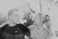 A black and white photograph of a smiling couple looking at each other, with the man's arm around the woman's shoulder. They are outdoors with a blurred background of trees.