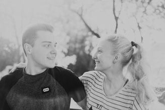A black and white photograph of a smiling couple looking at each other, with the man's arm around the woman's shoulder. They are outdoors with a blurred background of trees.