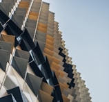 A modern architectural structure features a repetitive, geometric facade with angled panels creating a dynamic, textured surface. The panels are arranged in a gradient pattern from darker to lighter tones, providing depth and a sense of movement.