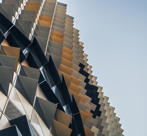 A modern architectural structure features a repetitive, geometric facade with angled panels creating a dynamic, textured surface. The panels are arranged in a gradient pattern from darker to lighter tones, providing depth and a sense of movement.