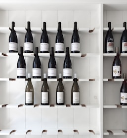 A minimalist display of wine bottles arranged on white wooden shelves, featuring predominantly dark glass bottles with a few lighter-colored ones. The labels on the bottles vary, with some showcasing simple text and others with more decorative designs.