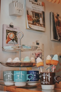 A cozy corner of a coffee shop featuring a tiered dessert stand with various cupcakes topped with colorful frosting. Each tier displays a different selection, including cupcakes with white, green, and orange frosting. Below, several mugs in different colors, labeled 'On the Way Coffee Shop', hold small marshmallow-like treats on sticks. In the background, several coffee-themed books and magazines are neatly mounted on the wall.
