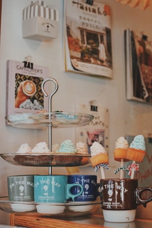 A cozy corner of a coffee shop featuring a tiered dessert stand with various cupcakes topped with colorful frosting. Each tier displays a different selection, including cupcakes with white, green, and orange frosting. Below, several mugs in different colors, labeled 'On the Way Coffee Shop', hold small marshmallow-like treats on sticks. In the background, several coffee-themed books and magazines are neatly mounted on the wall.