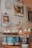 A cozy corner of a coffee shop featuring a tiered dessert stand with various cupcakes topped with colorful frosting. Each tier displays a different selection, including cupcakes with white, green, and orange frosting. Below, several mugs in different colors, labeled 'On the Way Coffee Shop', hold small marshmallow-like treats on sticks. In the background, several coffee-themed books and magazines are neatly mounted on the wall.