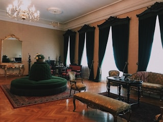 Elegant, vintage-style living room featuring a large chandelier, tall dark curtains, and intricate furniture pieces. A circular green cushioned seat is centrally placed on a decorative carpet. Ornate furniture such as a mirror, chairs, and a sofa are intricately designed, complementing the sophisticated ambiance.
