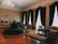 Elegant, vintage-style living room featuring a large chandelier, tall dark curtains, and intricate furniture pieces. A circular green cushioned seat is centrally placed on a decorative carpet. Ornate furniture such as a mirror, chairs, and a sofa are intricately designed, complementing the sophisticated ambiance.