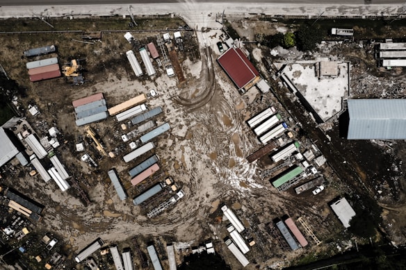 An aerial view of an industrial area with multiple rectangular buildings and structures, some with colorful roofs. The ground shows signs of construction activity and is muddy with tire tracks and scattered materials. The layout includes large vehicles and equipment, likely related to construction or transportation.