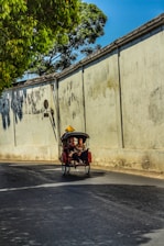 A person is sitting in a pedicab, traveling along a quiet street lined with a large, weathered wall. A leafy tree branch partially shades the street, and the sky is clear and blue.