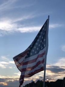 An American flag is waving on a flagpole against a backdrop of a partly cloudy sky at sunset. The flag is partially illuminated by the soft, golden sunlight, creating a serene and patriotic scene.