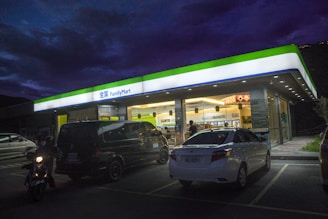 A brightly lit FamilyMart convenience store is prominent against a dark evening sky, with neon signage and a well-illuminated interior. Several vehicles, including a dark van and a white sedan, are parked in front of the store. A person on a scooter is approaching from the left, and another individual is visible inside the store near the entrance.