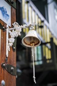 A rustic, metal bell with a white and rusted patina hangs on an intricately decorated metal bracket featuring floral designs. It is mounted on a wooden surface with visible metal fixtures. In the background, there is a blurred view of a balcony railing painted in dark colors with yellow highlights.