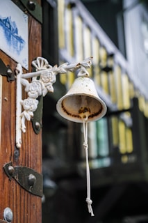 A rustic, metal bell with a white and rusted patina hangs on an intricately decorated metal bracket featuring floral designs. It is mounted on a wooden surface with visible metal fixtures. In the background, there is a blurred view of a balcony railing painted in dark colors with yellow highlights.