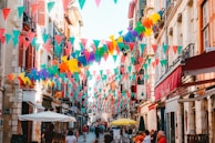 Colorful triangular flags hang across a narrow European street lined with historic buildings. People casually walk along, and a few tables with umbrellas are set up on the street. The sunlight casts warm tones on the facades, adding a vibrant atmosphere.