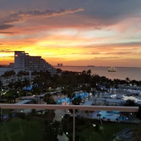 A scenic seaside view during sunset with a vibrant orange and yellow sky. A multi-story hotel building with a visible logo sits on the left side. Several swimming pools and palm trees are in the foreground, creating a tropical atmosphere. In the distance, a ship with visible lights is sailing on the water.