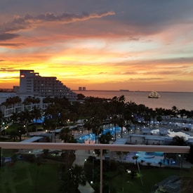 A scenic seaside view during sunset with a vibrant orange and yellow sky. A multi-story hotel building with a visible logo sits on the left side. Several swimming pools and palm trees are in the foreground, creating a tropical atmosphere. In the distance, a ship with visible lights is sailing on the water.