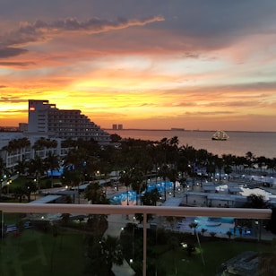 A scenic seaside view during sunset with a vibrant orange and yellow sky. A multi-story hotel building with a visible logo sits on the left side. Several swimming pools and palm trees are in the foreground, creating a tropical atmosphere. In the distance, a ship with visible lights is sailing on the water.