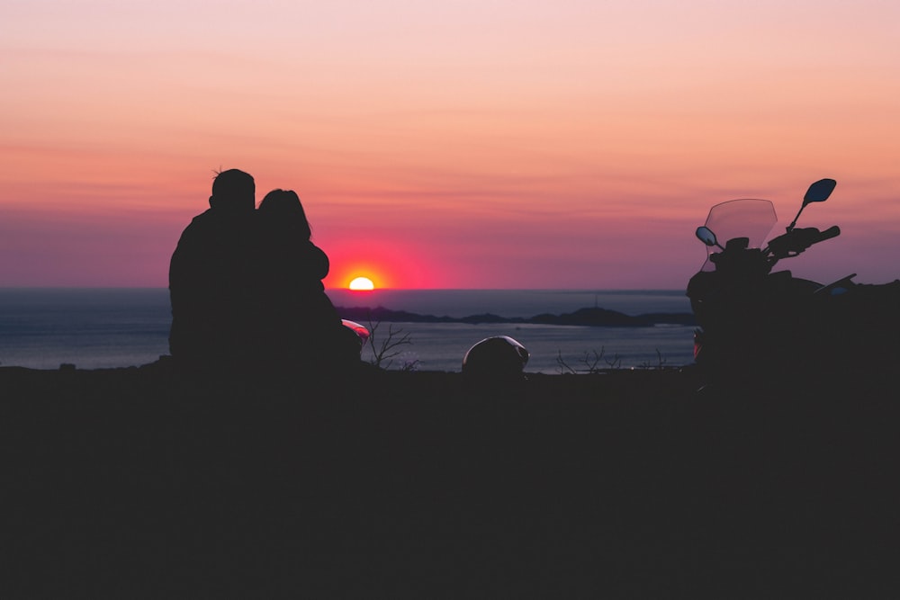 silhouette photo of man and woman sitting on grass beside motorcycle during golden hour