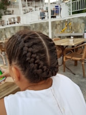 A young girl with neatly braided hair sits at an outdoor café. She is wearing a white top and holding a smartphone. The background shows a stone wall, wooden furniture, and some decorative elements like potted plants.