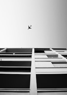 A black-and-white photograph capturing an upward view of a modern building facade with horizontal lines and an airplane flying overhead in the clear sky.