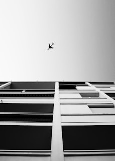A black-and-white photograph capturing an upward view of a modern building facade with horizontal lines and an airplane flying overhead in the clear sky.