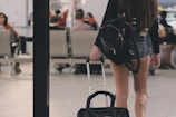 A person with long hair is seen from behind carrying a large black backpack and pulling a wheeled suitcase. They are wearing a black long-sleeved top and denim shorts. The setting is an airport or a similar transportation hub, with multiple people sitting in the background and a calm ambiance.