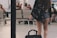 A person with long hair is seen from behind carrying a large black backpack and pulling a wheeled suitcase. They are wearing a black long-sleeved top and denim shorts. The setting is an airport or a similar transportation hub, with multiple people sitting in the background and a calm ambiance.