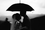 A silhouette of a couple sharing an intimate moment under an umbrella, with a mountainous backdrop. The woman appears to be wearing a veil, suggesting a wedding or romantic context.