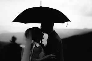 A silhouette of a couple sharing an intimate moment under an umbrella, with a mountainous backdrop. The woman appears to be wearing a veil, suggesting a wedding or romantic context.