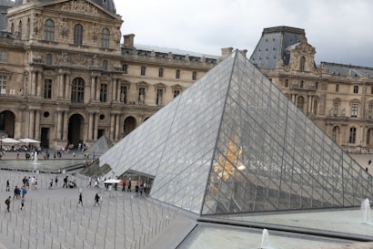 A large glass pyramid stands in the foreground with people walking around it. The background features an ornate, historic building with intricate architectural details and sculptures. The scene is bustling with tourists, suggesting a popular attraction.