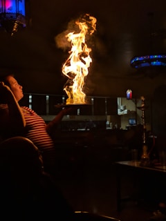 A person holds a tray with a large, active flame rising from it. The setting appears to be a dimly lit restaurant or bar, with a kitchen visible in the background. Decorative lamps with blue and red hues provide subtle lighting. There is a hookah pipe on a nearby table.
