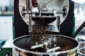 Close-up of a coffee roasting machine in action, with freshly roasted coffee beans pouring out into a metal tray. The machine has various metal components, knobs, and parts that help in the roasting process.