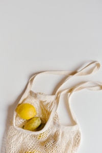 A white mesh reusable grocery bag contains a yellow lemon and some prickly pear fruits, placed on a plain light surface.