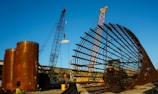 A large construction site featuring two cranes and a massive cylindrical metal structure. The site includes large rust-colored cylindrical vessels and a lattice framework of metal bars. The sky is clear and blue, adding contrast to the industrial elements.