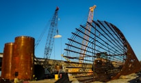 A large construction site featuring two cranes and a massive cylindrical metal structure. The site includes large rust-colored cylindrical vessels and a lattice framework of metal bars. The sky is clear and blue, adding contrast to the industrial elements.