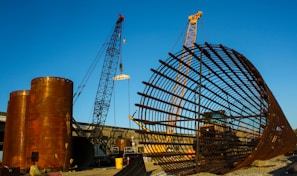 A large construction site featuring two cranes and a massive cylindrical metal structure. The site includes large rust-colored cylindrical vessels and a lattice framework of metal bars. The sky is clear and blue, adding contrast to the industrial elements.