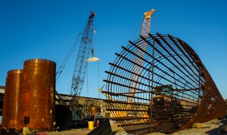 A large construction site featuring two cranes and a massive cylindrical metal structure. The site includes large rust-colored cylindrical vessels and a lattice framework of metal bars. The sky is clear and blue, adding contrast to the industrial elements.
