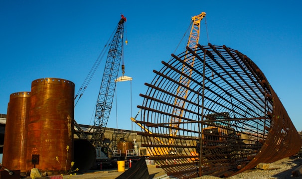 A large construction site featuring two cranes and a massive cylindrical metal structure. The site includes large rust-colored cylindrical vessels and a lattice framework of metal bars. The sky is clear and blue, adding contrast to the industrial elements.