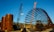 A large construction site featuring two cranes and a massive cylindrical metal structure. The site includes large rust-colored cylindrical vessels and a lattice framework of metal bars. The sky is clear and blue, adding contrast to the industrial elements.