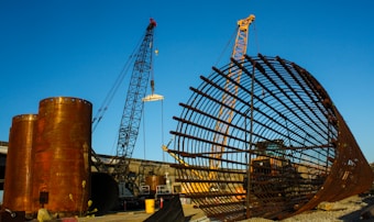 A large construction site featuring two cranes and a massive cylindrical metal structure. The site includes large rust-colored cylindrical vessels and a lattice framework of metal bars. The sky is clear and blue, adding contrast to the industrial elements.