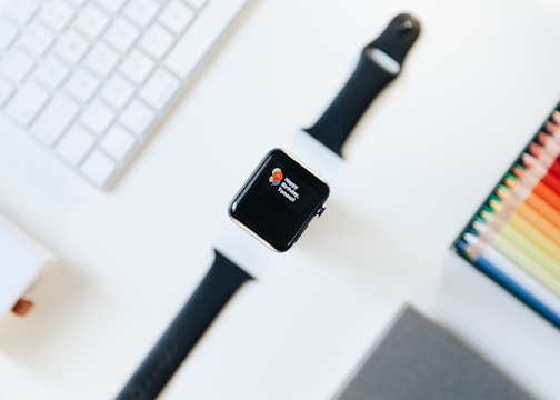 A smartwatch is centered on a desk, displaying a notification on its screen. Surrounding objects include a keyboard, a watch strap, and a set of colored pencils creating a vibrant, organized workspace.