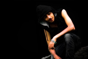 A young individual wearing a sleeveless black hoodie and jeans is seated against a dark background. The hood is up, partially covering the head, and a silver watch adorns the left wrist. The person's pose is relaxed with elbows resting on the knees.